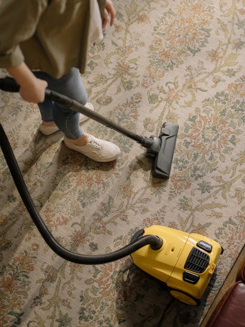A person operating a yellow portable carpet steam cleaner, focusing on the control panel, in a residential living room with a patterned rug, a brick wall in the background, and natural lighting. The i